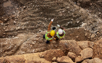 Two construction workers in high-visibility jackets and helmets inspecting a rocky excavation site, using a tablet and pointing at the terrain.