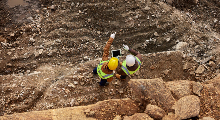 Two construction workers in high-visibility jackets and helmets inspecting a rocky excavation site, using a tablet and pointing at the terrain.