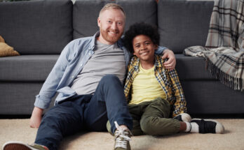 A smiling foster parent sitting on the floor with their happy foster child, sharing a warm moment in a cozy home environment.