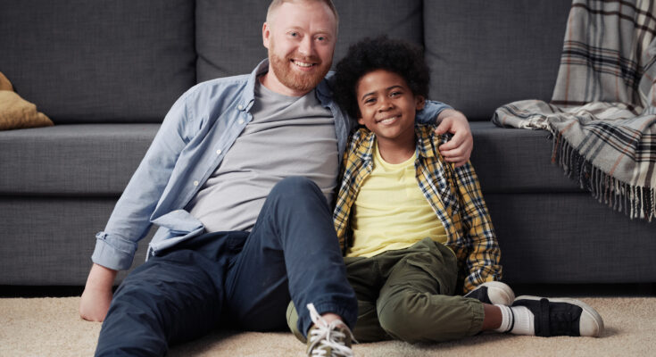 A smiling foster parent sitting on the floor with their happy foster child, sharing a warm moment in a cozy home environment.