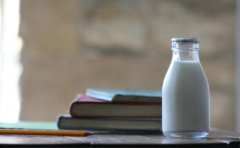 A small glass bottle of milk with a foil cap placed on a wooden surface, next to a stack of vintage books and a yellow pencil. The background is softly blurred, creating a warm, rustic atmosphere.
