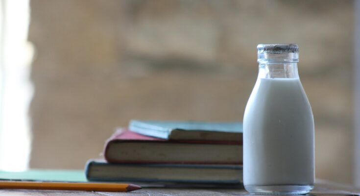 A small glass bottle of milk with a foil cap placed on a wooden surface, next to a stack of vintage books and a yellow pencil. The background is softly blurred, creating a warm, rustic atmosphere.